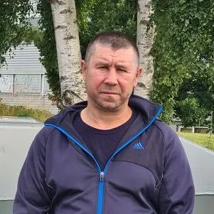 Mann i blå jakke står ute foran trær og hus